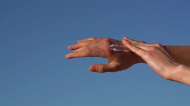 Bir kadının eli önce cildine besleyici bir krem sürer ve sonra onu mavi gökyüzü arka planına sürter. Cildi nemlendirme ve besleme kavramı, vücut bakımı için doğal kozmetik..