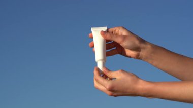 Kadın elleri, mavi gökyüzü arka planında beyaz bir model tüpten krem ya da serum sürerler. Güzellik ve estetik bakım ürünleri, yüz ve vücudun nemlendirilmesi ve beslenmesi kavramı.