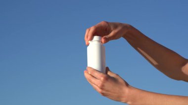 Kadın elleri beyaz bir ilaç şişesini açar ve mavi gökyüzü arka planına karşı avucunun içine hapları döker. Eczane ve ilaç kavramı, sağlık hizmetleri, vitaminler ve diyet takviyeleri