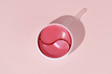 Pembe hidrojel göz bantları ve pembe izole edilmiş arka planda spatulası olan kavanozun üst görüntüsü. Doğal nemlendirici güzellik ürünleri kavramı, kırışıklık giderme, yüz gerdirme, koyu halkalar küçültme