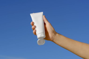 Kadın eli, mavi arka planda beyaz krem tüpünü tutuyor. Kozmetik kavramı, vücut ve yüz bakımı için doğal kozmetik. Tasarımın için resim