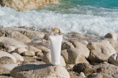 Sahildeki deniz suyunun arka planına karşı çakıl taşı taşının üzerindeki bir tüp kremin beyaz modeli. Doğal güzellik ürünleri, cilt bakımı, beslenme ve su kaybı, güneş koruması.