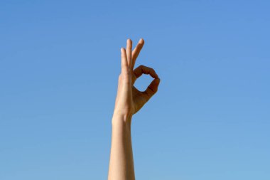Bir kadının elinin parmakları tamam ya da tamam işareti oluşturur. Bu da onay, rıza ve mavi gökyüzü arka planında her şeyin yolunda olduğuna dair bir işaret demektir..