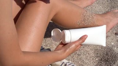 Genç bir kadın beyaz bir model tüpten eline krem sürüyor ve arka plandaki kumlara bacağına sürtünüyor. Vücudu güneşten, cilt bakımından ve nemlendiriciden, güzellik ürünlerinden, dermatolojiden korumak için kullanılan güneş kremi kavramı..