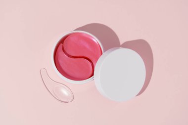 Pembe hidrojel yamalı beyaz maket kavanozu ve pembe izole edilmiş arka planda şeffaf spatula. Nemlendirici ve besleyici yüz derisi konsepti, güzellik ve estetik ürünleri