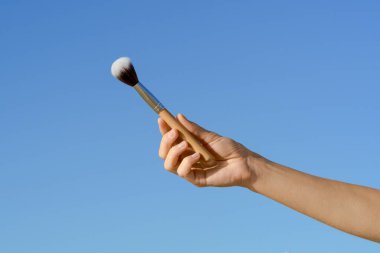 Mavi izole gökyüzü arka planında yüz makyajı için doğal ahşap fırçayı tutan genç bir kadının eli. Dekoratif kozmetik kavramı, yüz bakımı, yüz bakımı