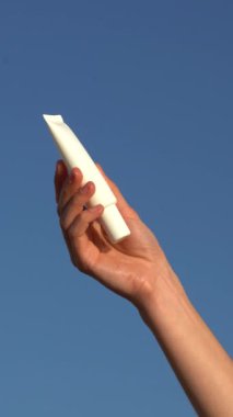Mavi bir arka planda beyaz bir krem tüpü ya da serum tutan bir kadının dikey videosu. Cildi nemlendirme ve besleme kavramı, yüz bakımı için doğal kozmetik..