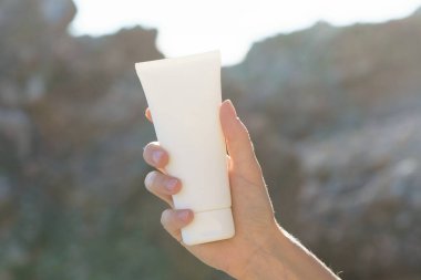 Genç bir kadının eli, deniz kenarındaki bir kaya zemininde güneş tarafından aydınlatılmış beyaz bir krem tüpünü tutuyor. Saç, yüz ve vücut için doğal kozmetik kavramı, güneş koruması, güzellik ürünleri.