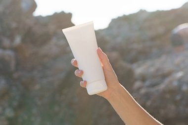 Genç bir kadının eli kayanın arka planında güneş tarafından aydınlatılmış beyaz bir krem tüpünü tutuyor. Güzellik ürünleri, cilt koruma ve bakım kavramı, doğal nemlendirici..