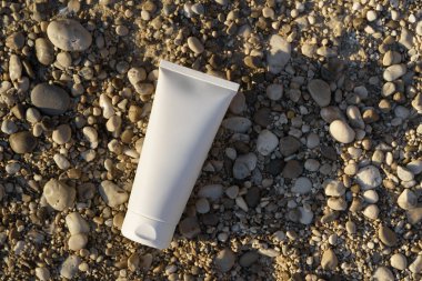 Kumsala yakın bir çakıl taşının üzerinde duran beyaz krem tüpü ve güneş ışığı. Doğal güzellik ürünleri, vücut ve yüz bakımı, güzellik ve estetik