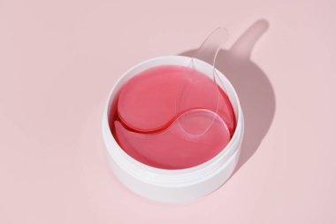 Pembe izole edilmiş arka planda pembe hidrojel göz bantları tutan parlak beyaz bir kavanoz. Deri hidrasyonunu, pürüzleri, kırışıklıkları arttırır ve koyu halkalar kavramını aydınlatır. Tasarımın için resim