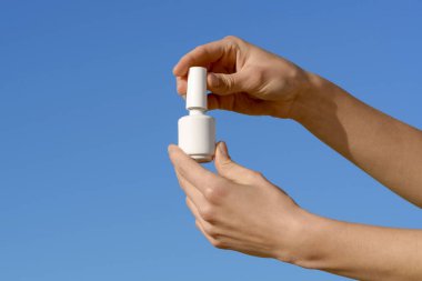 Mavi izole bir arka planda beyaz bir tırnak cilası kavanozunu tutan ve açan kadın elleri. Güzellik ürünleri, jel cilası, güzellik salonları ve estetik.