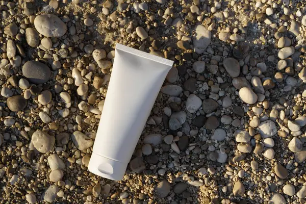 Kumsala yakın bir çakıl taşının üzerinde duran beyaz krem tüpü ve güneş ışığı. Doğal güzellik ürünleri, vücut ve yüz bakımı, güzellik ve estetik