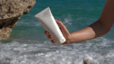 Genç bir kadının eli sahildeki deniz dalgalarının arka planında beyaz bir krema tüpü tutuyor. Cilt, yaz bakımı ve nemlendirici güneş kremi ürünleri kavramı