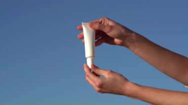 Dişi eller, beyaz bir model tüpü serum ya da krem ile açar ve mavi gökyüzü arka planındaki cilde uygular. Güzellik ve yüz bakımı ürünleri kavramı, güzellik ve estetik ürünleri, spa, kozmetoloji.