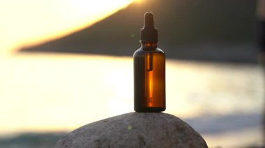 Gün batımı denizi ve dalgalanan suyun arka planına karşı bir taşın üzerinde şeffaf cam kahverengi serum kavanozu. Doğal cilt bakım ürünleri, güzellik ürünleri kavramı