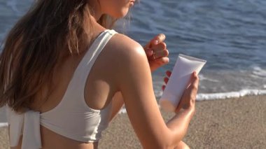 Mayo giymiş, bol saçlı genç bir kadın beyaz bir model tüp tutuyor ve omzuna plajdaki deniz dalgalarının arka planına güneş kremi sürüyor. Cilt bakımı ve koruma kavramı