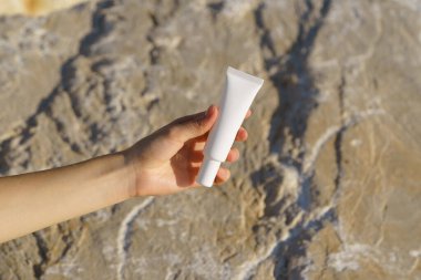 Kadının eli, deniz taşının arka planına karşı beyaz bir krem tüpü tutuyor. SPF güneş kremi konsepti, cilt bakımı, doğal güzellik kozmetiği