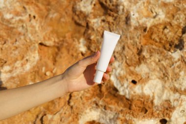 Deniz kenarındaki kireçtaşı zemininde ince beyaz bir göz kremi tüpü ya da serum tutan bir kadının yakın çekimi. Cilt bakımı, nemlendirici, beslenme, güneş kremi, yaşlanmayı önleme.