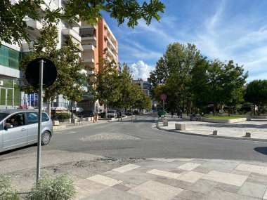 Vlora, Arnavutluk - 16 Ocak 2025: Arnavutluk 'ta arabaları, yeşil çalıları ve binaları olan güneşli bir cadde. Akdeniz günlük yaşamının ve bölgesel kentsel cazibenin manzaralı bir temsili.