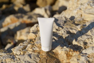 Güneş ışığı altında kayalık bir yüzeye yerleştirilmiş beyaz makyaj tüpü. Sahne, cilt bakımı ya da güzellik bağlamında ürünü ön plana çıkarıyor.