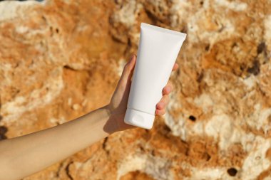 Denize yakın kireçtaşı bir zeminde beyaz bir krem tüpünü tutan bir kadının yakın çekimi. Cilt bakımı, nemlendirici, spa beslenmesi, güzellik ürünleri, güneş kremi kavramı.