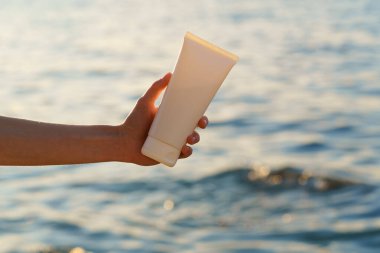 Batan güneşin aydınlattığı deniz dalgalarının arka planında beyaz bir krem tüpünü tutan kadın el. Kozmetik güzellik ürünleri, nemlendirici ve doğal cilt bakımı kavramı