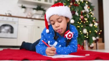 Mavi kazaklı ve şapkalı küçük bir Afro-Amerikalı Noel ağacının arkasına Noel Baba 'ya bir dilek mektubu yazıyor..