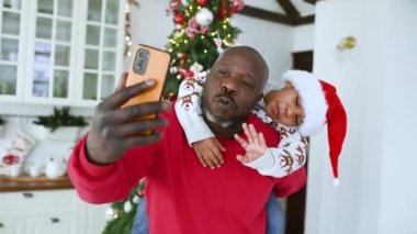 Kırmızı kazaklı bir baba, Noel Baba şapkası takmış küçük bir oğlu var. Bir yılbaşı ağacının arka planında kendini kamerayla çekerken gülümsüyor..