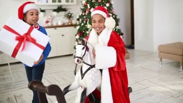 サンタの衣装を着た弟と妹と鹿とセーター ロッキングホースに乗って 手にクリスマスプレゼント付きの巨大な箱を持っています — ストック動画