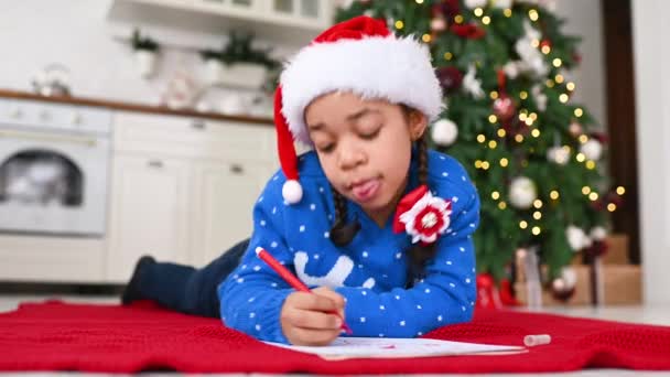 一个身穿蓝色毛衣 头戴帽子的非洲裔美国小女孩在圣诞树下给圣诞老人写了一封信 — 图库视频影像