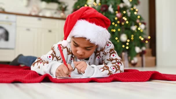 一个头戴圣诞礼帽 头戴圣诞礼帽的小男孩梦幻般地微笑着 并在一张白色的纸片上给圣诞老人写了一封信 一张挂在圣诞树下的愿望清单 — 图库视频影像