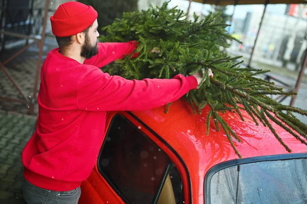 a young man in a red sweater and a red hat puts a Christmas tree on the roof of a red retro car against the background of falling snow.