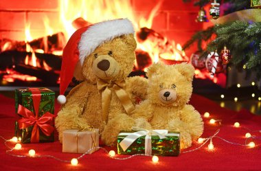 Noel Baba şapkalı iki oyuncak ayı, hediyelerin ve yılbaşı ışıklarının önünde sıcacık bir şöminenin önünde birbirlerine sarılıyorlar.