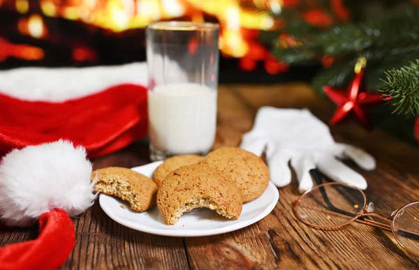 a bitten oatmeal cookie and an unfinished glass of milk on a wooden table against the background of Santa Claus clothes, a Christmas tree and a fireplace