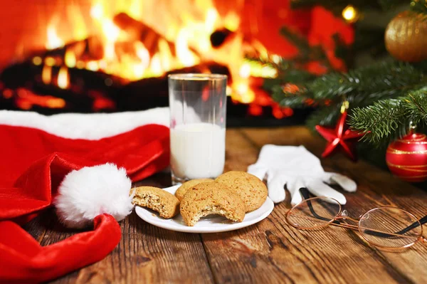 bitten oatmeal cookie and an unfinished glass of milk on a wooden table against the background of Santa Claus clothes a Christmas tree and a fireplace