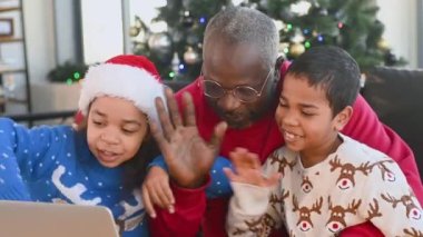 Afrika kökenli Amerikan ailesi - Noel Baba, oğul ve kız Noel Baba kazakları ve Noel Baba şapkalı kucaklaşma, gülümseme ve Noel ağacının arka planındaki akrabalarla video bağlantısı yoluyla iletişim.