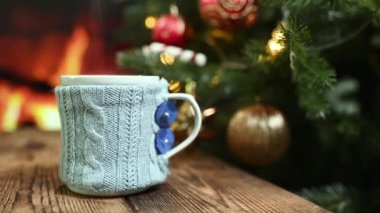 Yanan bir şöminenin ve Noel ağacının arka planında, örgü bir çantanın içinde sıcak bir içecek ve ahşap bir masanın üzerinde bir kupa.