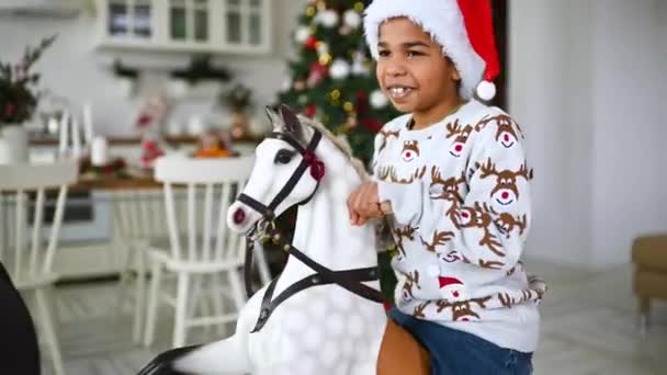 一个身穿灰色圣诞毛衣 头戴红色桑塔帽的非洲裔小男孩坐在圣诞树和节庆桌旁摇曳的玩具马背上笑了 — 图库视频影像