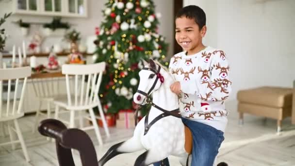 クリスマスツリーとお祝いのテーブルを背景におもちゃのロッキングホースに座っている間 鹿の笑顔で灰色のクリスマスセーターの小さなアフリカ系アメリカ人の少年 — ストック動画