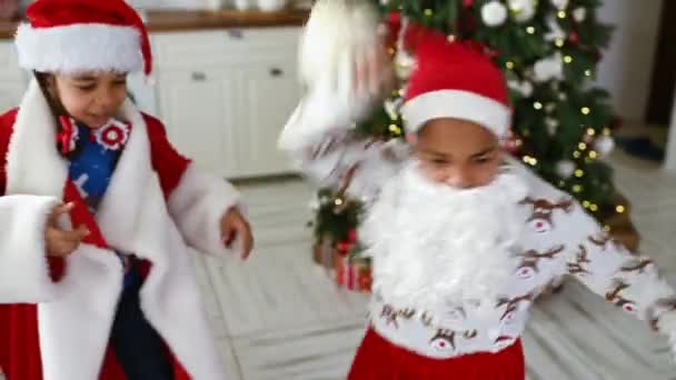 小さな子供たち男の子と女の子はサンタクロースの格好をしたクリスマスツリーの背景に楽しいとダンスを持っています — ストック動画