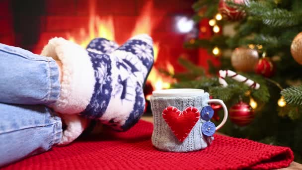 脚穿保暖袜子 有斯堪的纳维亚装饰 背靠壁炉 圣诞树 格子花和装在心形针织被子里的杯子 — 图库视频影像