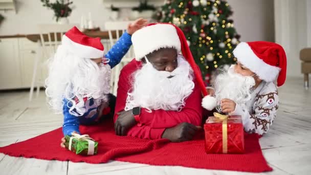 アフリカ系アメリカ人の家族 サンタの衣装を着た父 そして彼らの手の中の贈り物は クリスマスツリーと装飾を背景にした赤い遊びと笑顔にあります — ストック動画