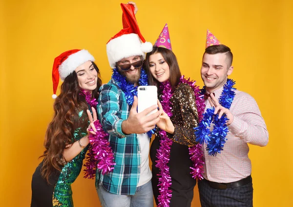 a group or company of friends in santa hat, party hats, boas celebrating Christmas, having fun, taking selfies and going live on a yellow background.