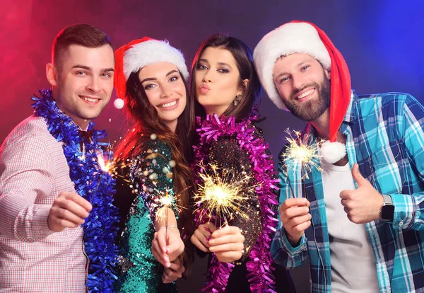 a group or company of friends in a santa hat and colored boas celebrating Christmas and smiling holding sparklers in their hands.
