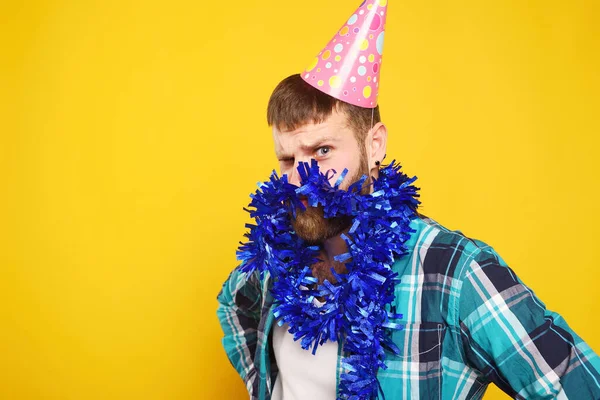 funny bearded man in a pink party cap with a blue boa on his beard grimaces on a yellow background