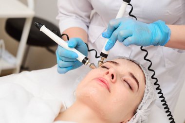 A cosmetologist conducts microcurrent facial therapy for a young woman using a device in a beauty salon. Hardware cosmetology, skin care, rejuvenation, regeneration.