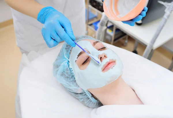 A cosmetologist puts an alginate mask on the face of a young pretty woman. Facial skin care, regeneration, rejuvenation