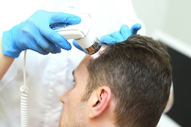 Trichoscopy is a computer examination of the scalp and hair. A cosmetologist-trichologist diagnoses the condition of a male patients hair with gray hair using a trichoscope.
