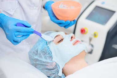 A cosmetologist puts an alginate mask on the face of a young pretty woman. Facial skin care, regeneration, rejuvenation